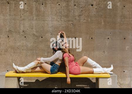 Zwei modische junge Frauen in trendigen Outfits entspannen sich und posieren auf einer hellgelben Bank vor einem strukturierten Betonhintergrund, um das Sonnenlicht zu genießen Stockfoto