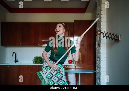 Ein verspieltes Bild einer kaukasischen Frau, die in Vintage-Kleidung der 50er Jahre gekleidet ist und freudig einen Besenstiel in ihrer Küche hält, während sie wegschaut, in eine grüne tu gekleidet Stockfoto