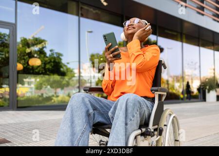 Eine fröhliche Afroamerikanerin im Rollstuhl, trägt orangefarbenes Oberteil und Jeans, benutzt ihr Handy an einem sonnigen Tag vor einem modernen Gebäude Stockfoto