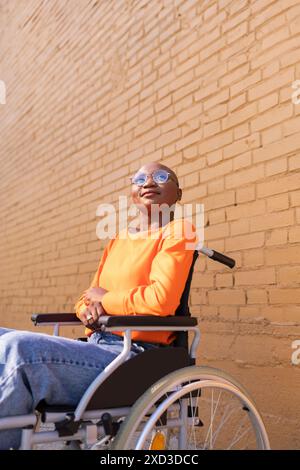 Eine Afroamerikanerin mit Sonnenbrille und orangefarbenem Oberteil sitzt souverän in einem Rollstuhl vor einer strukturierten Ziegelwand, im Sonnenlicht getaucht Stockfoto