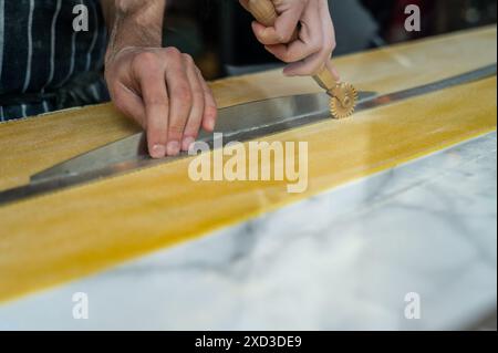 Nahaufnahme eines zugeschnittenen, nicht erkennbaren Küchenchefs, der frischen Teig mit einem traditionellen Nudelschneider ausrollt. Stockfoto