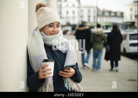 Modisch gekleidete junge Frau genießt ihren Kaffee, während sie ihr Smartphone auf einer belebten Londoner Straße überprüft. Stockfoto