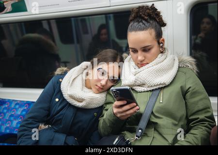 Zwei junge Frauen, gebündelt in warmer Kleidung und Schals, konzentrieren sich auf ein Smartphone, während sie in einer Londoner U-Bahn sitzen und einen Moment der Moderne reflektieren Stockfoto