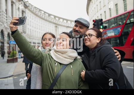 Eine fröhliche vierköpfige Familie, darunter zwei Töchter mit ihren Eltern, lächelt, während sie in London ein Selfie mit einem Smartphone machen, mit einem roten Bus und Stockfoto