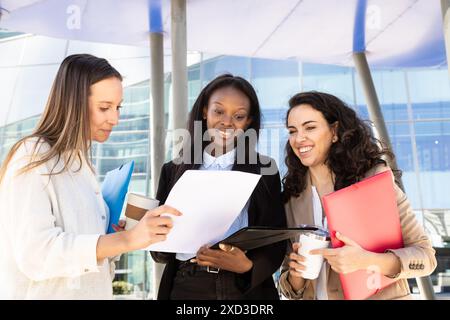 Drei multirassische Frauen mit unterschiedlichem ethnischen Hintergrund führten eine lebhafte Diskussion mit Dokumenten und Kaffee in der Hand, vor einem modernen Glas-bui Stockfoto