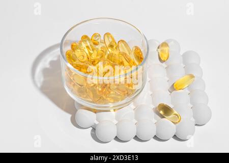 Transparente Schüssel gefüllt mit goldenen Omega-3-Kapseln mit verstreuten weißen Pillen rund um den weißen Hintergrund ideal für Gesundheits-Ergänzungsaktionen Stockfoto