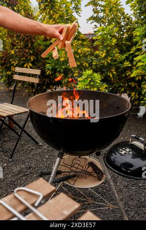 Beschnittene, nicht erkennbare Person, die einen Holzkohlegrill mit Holzstöcken in einem Hinterhof in Brand setzt. Stockfoto