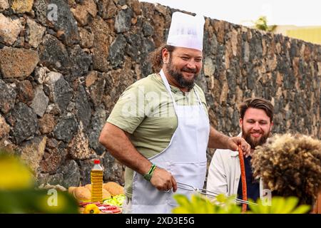Bei einem Treffen im Freien grillt ein fröhlicher Mann in einem Hut des Küchenchefs, umgeben von Freunden, die Musik genießen. Das Setting umfasst verschiedene ethnische Zugehörigkeiten in Stockfoto