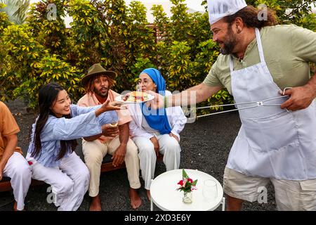 Ein Mann serviert Speisen für eine Gruppe von Freunden bei einem BBQ im Hinterhof. Eine asiatische Frau, ein afroamerikanischer Mann und eine hispanische Frau im Hijab engagieren sich fröhlich Stockfoto