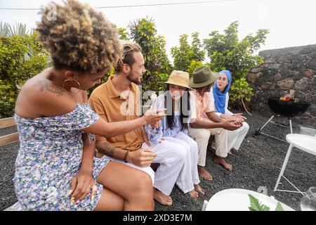 Eine vielfältige Gruppe von Freunden, darunter afroamerikanische, kaukasische, asiatische und nahöstliche Personen, verbringen einen Moment bei einem Grillabend im Hinterhof Stockfoto