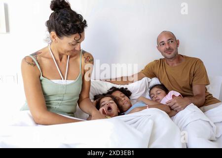 Eine multiethnische Familie genießt einen gemeinsamen Morgen im Bett, zwei Kinder wachen zwischen ihren lächelnden Eltern auf und zeigen eine Szene voller Wärme Stockfoto