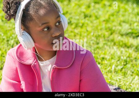 Ein junges schwarzes Mädchen sitzt in einem üppigen Park und genießt Musik über ihre weißen Kopfhörer Sie trägt eine leuchtend rosa Jacke über einem weißen Hemd Stockfoto