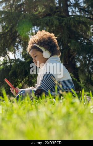 Ein junges Mädchen sitzt auf dem Gras in einem Park, während es über weiße Kopfhörer in ihre Musik eingetaucht ist, während es ein rotes Smartphone benutzt Stockfoto