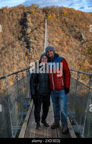 Ein fröhliches Paar, das auf der Geierlay-Hängebrücke in Deutschland steht, umgeben von herbstfarbenen Wäldern, die die Brücke zeigen.“ Stockfoto