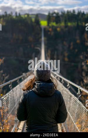 Rückansicht einer nicht erkennbaren Frau, die auf der Geierlay-Hängebrücke steht und auf den Weg vor Ihnen blickt, umgeben von Herbstlaub. Diese heitere sce Stockfoto