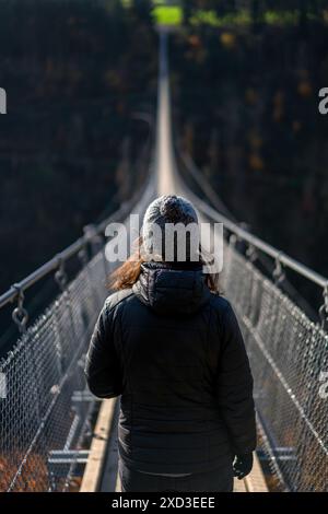 Rückansicht einer nicht erkennbaren Frau mit Winterjacke und Mütze, die über die berühmte Geierlay-Hängebrücke in Deutschland läuft. Die Brücke s Stockfoto