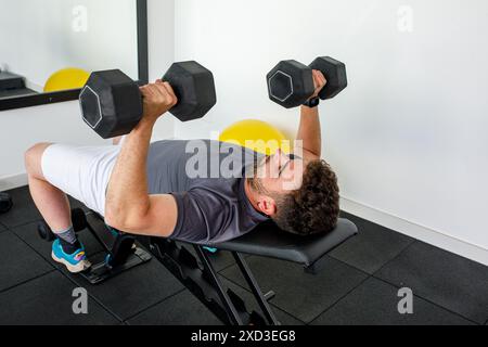 Ein fokussierter Mann drückt mit schweren Kurzhanteln auf der Bank, während er auf einer Turnbank liegt, umgeben von minimalistischen Fitnessgeräten und einem hellen Innenraum Stockfoto