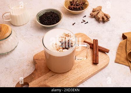 Von oben köstlicher Chai Latte mit cremigem Schaum auf einem Holzbrett umgeben von Sternanis, Zimtstangen, frischem Ingwer, Teeblättern, einem Milchglas und Stockfoto