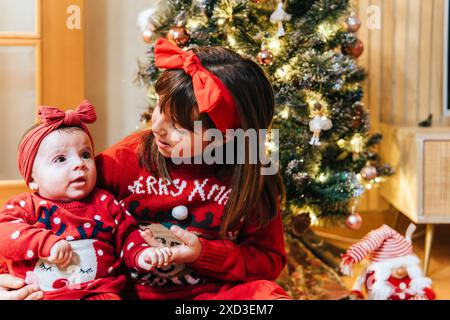 Ein junges Mädchen in einem festlichen Pullover schaut ihr Geschwister, ein Kleinkind in einem Weihnachts-Einteiler, mit einem geschmückten Baum im Hintergrund. Stockfoto