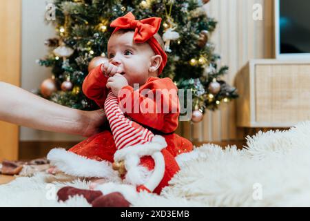 Kleinkind trägt ein rotes Weihnachtsmann-Outfit mit einer Schleife und blickt weg, während er an einem Weihnachtsbaum sitzt. Erwachsene, nicht erkennbare Armstütze. Stockfoto