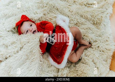 Von oben liegt der Blick auf ein kleines Mädchen in einem roten Weihnachtsoutfit mit Schleife auf einer flauschigen Decke Stockfoto