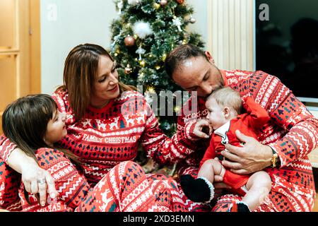 Eine vierköpfige Familie mit passendem rotem Family Christmas Pyjama sitzt neben ihrem geschmückten Weihnachtsbaum und hängt über einem Baby. Stockfoto