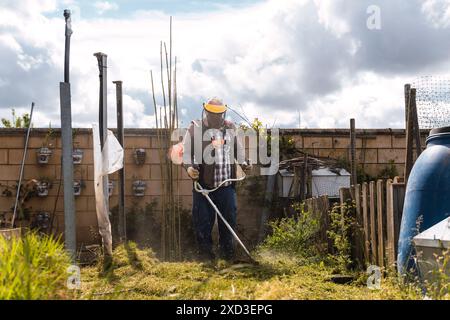 Ein Mann in Schutzkleidung verwendet einen Unkrautschneider, um einen bewachsenen Garten zu verwalten und an einem sonnigen Tag Themen wie Gartenarbeit und Pflege hervorzuheben Stockfoto