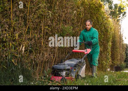 Ein Mann in grünen Overalls konzentriert sich darauf, das Gras mit einem rot-schwarzen Rasenmäher zu mähen, umgeben von üppigem Grün und Bambus Stockfoto