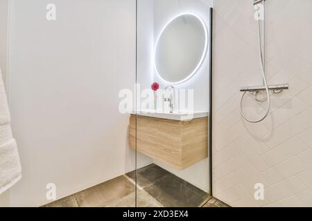 Ein modernes Badezimmer mit einem eleganten, an der Wand montierten Kosmetikbereich mit beleuchtetem runden Spiegel, einem einzelnen Blumenakzent und einer minimalistischen Dusche Stockfoto