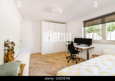 Ein gut beleuchtetes, modernes Schlafzimmer mit Home Office-Einrichtung, geräumigem Arbeitsplatz mit zwei Monitoren, Parkettboden und minimalistischer Einrichtung. Stockfoto