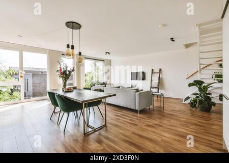 Die wunderschön beleuchtete Inneneinrichtung zeigt ein modernes Wohnzimmer mit dunkelgrünen Stühlen, einem eleganten Esstisch und elegantem Dekor. Die Fenster bieten viel natürliches li Stockfoto