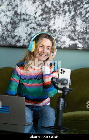 Eine fröhliche junge Frau mit Kopfhörervlogs von zu Hause, die intelligentes Arbeiten und Multimedia-Engagement zeigen Stockfoto