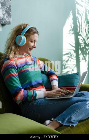 Eine junge Frau arbeitet gern zu Hause intelligent, verwendet einen Laptop und trägt Kopfhörer, sitzt bequem auf einem grünen Sofa mit Naturdarstellungen auf t Stockfoto