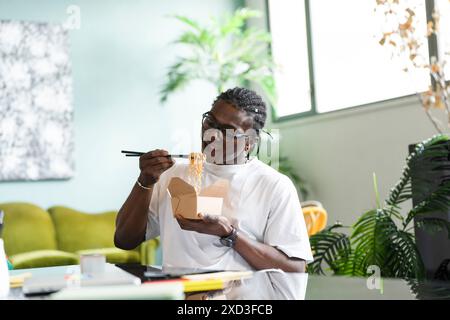 Ein schwarzer Mann sitzt in einem komfortablen Innenbereich und isst mit Essstäbchen Nudeln aus einer Take-Out-Box Stockfoto