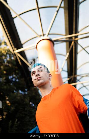 Ein stilvoller junger Mann mit auffälligen blauen Haaren und modernen Ohrringen steht selbstbewusst draußen, gekleidet in einem leuchtend orangen Hemd vor dem Hintergrund von ge Stockfoto