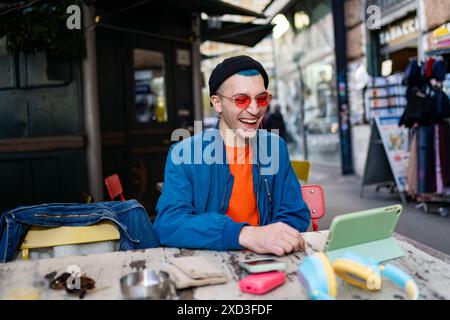 Ein fröhlicher junger Mann mit Mütze und roter Sonnenbrille lacht herzlich, während er an einem Café-Tisch sitzt, umgeben von seinem Smartphone und seinem Kopfhörer Stockfoto