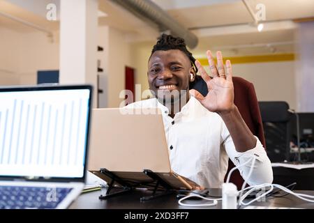 Ein dynamischer afroamerikanischer Mann in einem modernen Büro winkt fröhlich zu seiner Laptop-Kamera und signalisiert eine freundliche Begrüßung oder ein erfolgreiches virtuelles Treffen Stockfoto
