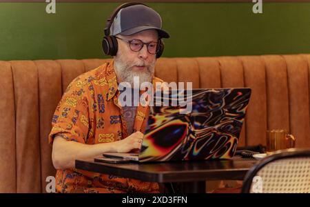 Ein moderner Senior mit stilvollem Bart arbeitet ferngesteuert an einem Laptop in einem Café, trägt Kopfhörer und konzentriert sich auf den Bildschirm inmitten von Naturillustrat Stockfoto