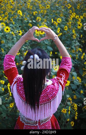 Eine junge Frau im Tribal-Outfit hebt ihre Hände, um ein herzförmiges Zeichen zu machen, das Liebe und Freundschaft vor dem Hintergrund eines wunderschönen Morgenflusses symbolisiert Stockfoto