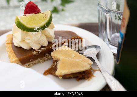 Nahaufnahme einer Scheibe Karamelltorte mit Schlagsahne, Limette und einer Himbeere, serviert auf einem weißen Teller mit einem Löffel. Stockfoto