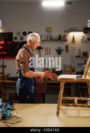 Konzept der Möbelreparatur, Heimwerkerarbeit und Heimpflege älterer Mann in der Werkstatt, der Handschuhe auflegt. Machen Sie sich bereit, alte wiederherzustellen Stockfoto