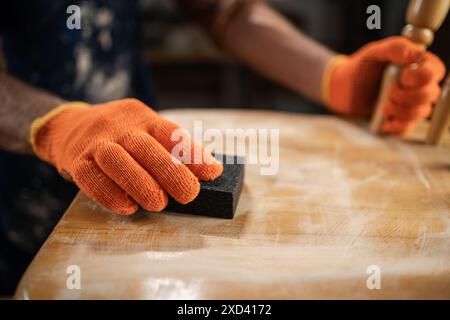 Möbelreparatur, Heimwerkerarbeit und Hausverbesserungskonzept. Mann in orangefarbenen Handschuhen, die Holzstuhl mit Schleifpapier schleifen, Oberfläche für die weitere Verarbeitung vorbereiten Stockfoto