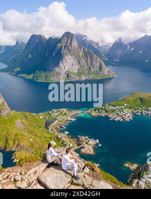 Ein Paar bewundert einen malerischen Fjord auf den Norways Lofoten Islands. Hoch aufragende Berge überblicken das kristallklare Wasser und ein malerisches Dorf darunter. Reinebringen, Lofoten, Norwegen Stockfoto