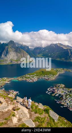 Ein Paar bewundert einen atemberaubenden Blick auf eine norwegische Küstenstadt zwischen zerklüfteten Bergen und strahlend blauem Wasser unter einem klaren blauen Himmel mit flauschigen weißen Wolken. Reinebringen, Lofoten, Norwegen Stockfoto