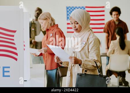Mittlere Aufnahme einer jungen muslimischen Frau im Hijab, die am Wahllokal steht und sich die Liste der Präsidentschaftskandidaten ansieht Stockfoto