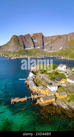 Ein Blick aus der Vogelperspektive auf ein kleines Dorf in Norwegen, mit farbenfrohen Häusern, die auf Stelzen über dem klaren blauen Wasser gebaut sind. Die Berge im Hintergrund bieten eine dramatische Kulisse. Sakrisoy, Lofoten, Norwegen Stockfoto