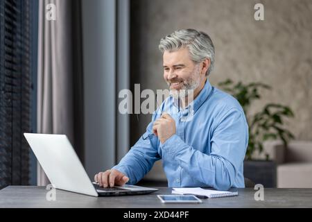 Senior-Mann, der am Laptop arbeitet, lächelt, während er in einem modernen Büro sitzt. Der Mann trägt ein blaues Hemd und scheint an seiner Arbeit beteiligt zu sein. Stockfoto