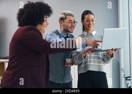 Drei Mitarbeiter im Büro, die sich den Laptop ansehen, sind überrascht von den wirtschaftlichen Vorteilen des Monats, und zeigen auf etwas auf dem Bildschirm. Sind zwei Frauen und Stockfoto