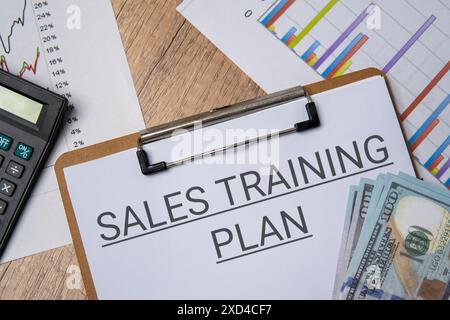 Draufsicht eines Clipboards mit den Worten „SALES TRAINING PLAN“ (VERKAUFSTRAINING PLAN) neben einem Stapel US-Dollar und einem Taschenrechner. Wirtschaft und Finanzen Stockfoto