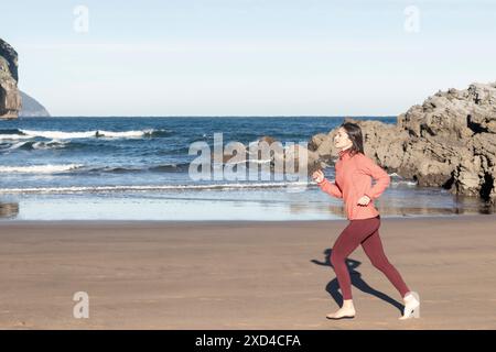 Junge Frau, die barfuß am Strand joggt in rosa Jacke und roter Leggings. Fitness- und gesunde Lifestyle-Fotografie. Sport- und Wellnesskonzept Stockfoto